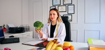 Female nutritionist in office holding broccoli, surrounded by fruits, promoting healthy lifestyle.