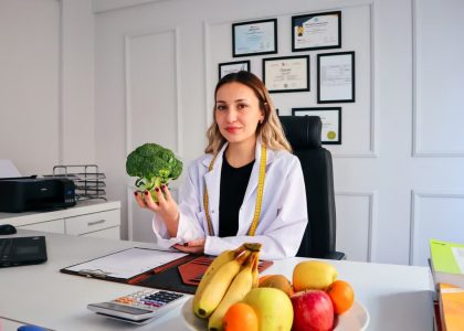 Female nutritionist in office holding broccoli, surrounded by fruits, promoting healthy lifestyle.